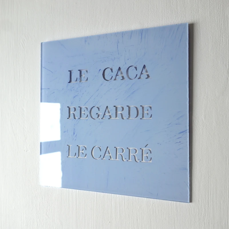 Un tableau carré en verre peint en bleu sur l'envers. La peinture est grattée pour écrire : le caca regarde le carré. Vue de trois-quarts.