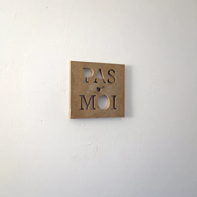 Une planche usagée est grossièrement découpée à la scie sauteuse pour former des lettres qui indiquent PAS MOI. L'ensemble est accroché à un clou dans le mur qui passe dans les lettres découpées.