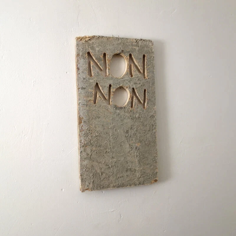 Une planche usagée est grossièrement découpée à la scie sauteuse pour former des lettres qui indiquent NON NON. L'ensemble est accroché à un clou dans le mur qui passe dans les lettres découpées.