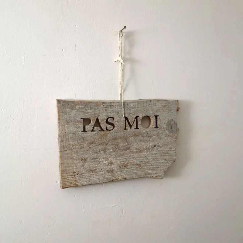 Une planche usagée est grossièrement découpée à la scie sauteuse pour former des lettres qui indiquent PAS MOI. L'ensemble est accroché à un clou dans le mur par une cordelette de récupération qui passe dans les lettres découpées.