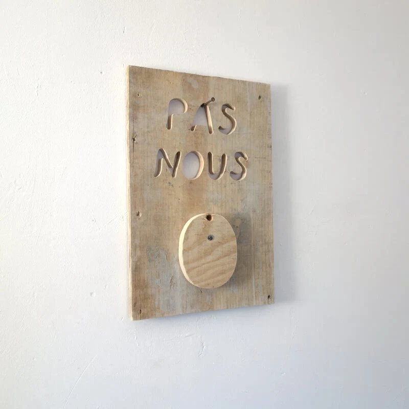 Une planche usagée est grossièrement découpée à la scie sauteuse pour former des lettres qui indiquent PAS NOUS. L'ensemble est accroché à un clou dans le mur qui passe dans les lettres découpées.