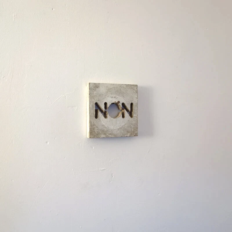 Une planche usagée est grossièrement découpée à la scie sauteuse pour former des lettres qui indiquent NON. L'ensemble est accroché à un clou dans le mur qui passe dans les lettres découpées.
