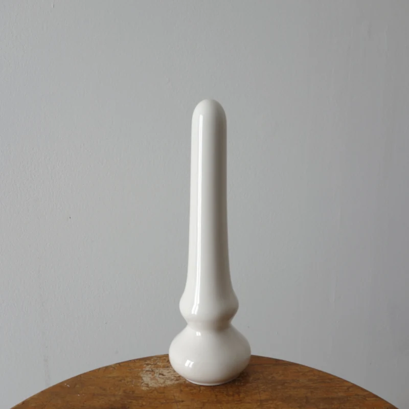 Un godemiché en céramique, blanc brillant, est posé sur un support en bois devant un fond indéterminé.